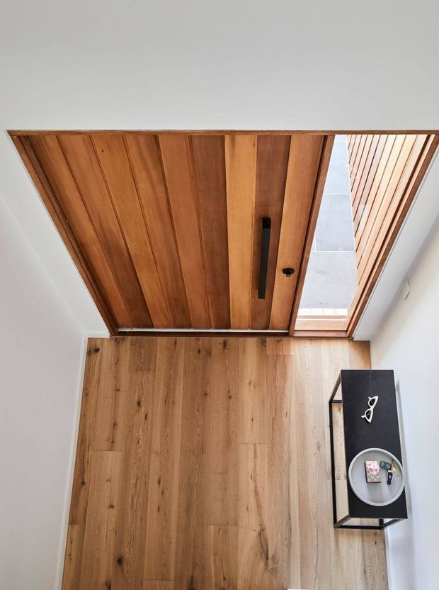 A compact entryway viewed from above, featuring a warm timber plank pivot or sliding door, matching light timber flooring, white walls, and a narrow black console table with a small tray and decorative object. A roof opening or glazed overhead section brings in natural light.