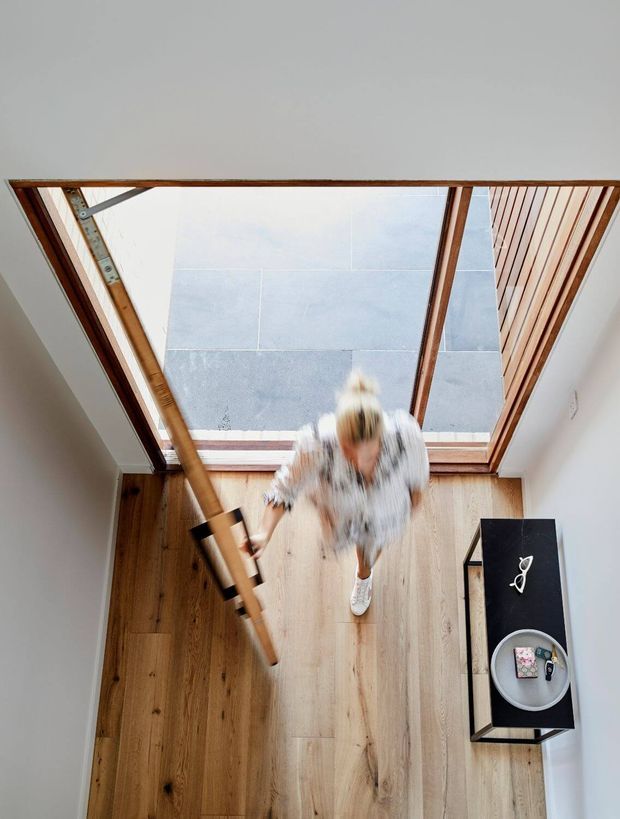 Overhead view of a bright contemporary interior with warm timber flooring, white walls, and a large timber-framed skylight or roof window. A blurred person stands beside a long suspended or leaning timber element, with a narrow black console table and bowl along the right wall.