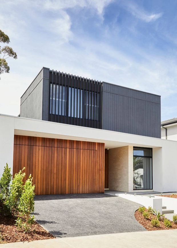 Front exterior view of a contemporary two-storey residence. The facade combines dark vertically profiled cladding and projecting vertical fins around upper-level glazing with a crisp white horizontal canopy, a warm timber garage door, pale masonry, and black-framed windows. A broad textured driveway and restrained low planting complete the street-facing composition.