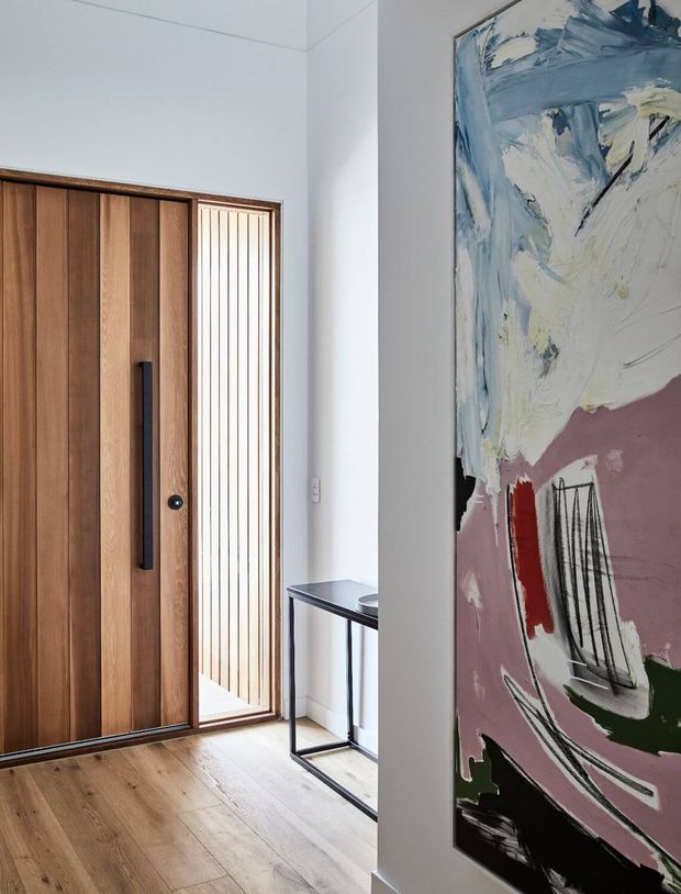 A bright contemporary entryway featuring a tall timber pivot front door with a slim black handle, a narrow vertical glazed sidelight with timber battens, light timber flooring, and a minimalist black metal console table. A large abstract artwork in blue, cream, mauve, red, green, and black occupies the wall in the foreground.