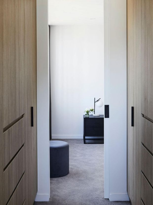View through a narrow hallway or wardrobe passage into a minimalist bedroom. Full-height light timber built-in cabinetry with dark recessed handles lines both sides, opening toward a softly carpeted room with a dark upholstered ottoman, black low dresser, table lamp and small plant. The palette is restrained in white, pale grey, light timber and black.