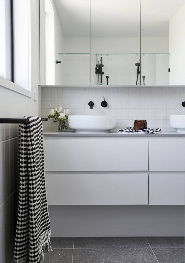 Bright contemporary bathroom featuring a floating white vanity with two vessel basins, a pale grey benchtop, white square-tile splashback, matte black tapware, large mirror, and a clear glass shower enclosure. Dark grey floor tiles and a black-and-white towel provide contrast, while a small vase and candle add styling.
