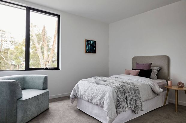 A bright, contemporary bedroom with a double bed, upholstered rounded headboard, layered white and grey bedding, a small timber bedside table, and a blue-grey armchair. A large black-framed window provides views of trees, while a small artwork adds a subtle accent to the otherwise neutral room.