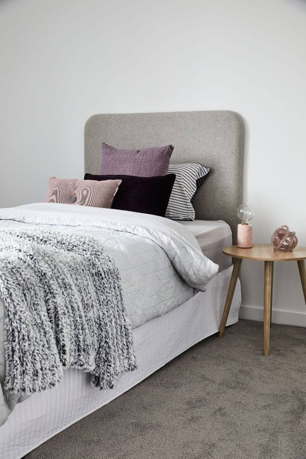 A softly styled bedroom with a large upholstered grey headboard, layered white bedding, textured grey throw, and assorted decorative pillows. A small round timber bedside table holds a lamp and decorative object. The room has pale walls, carpeted flooring, and a calm neutral palette with muted mauve accents.