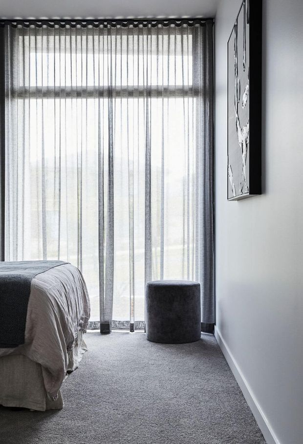 A minimalist bedroom corner with a partially visible upholstered bed, soft grey carpet, floor-to-ceiling sheer grey curtains over a large window, and a dark cylindrical ottoman. The room has white walls, subdued monochromatic furnishings, and framed black-and-white artwork, creating a calm, modern atmosphere.