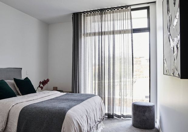 Bright contemporary bedroom with a neatly made bed, white bedding and a charcoal grey throw, dark teal accent cushions, sheer floor-to-ceiling curtains, and a tall black-framed window. A small grey cylindrical ottoman and monochrome wall artwork complete the restrained modern scheme.