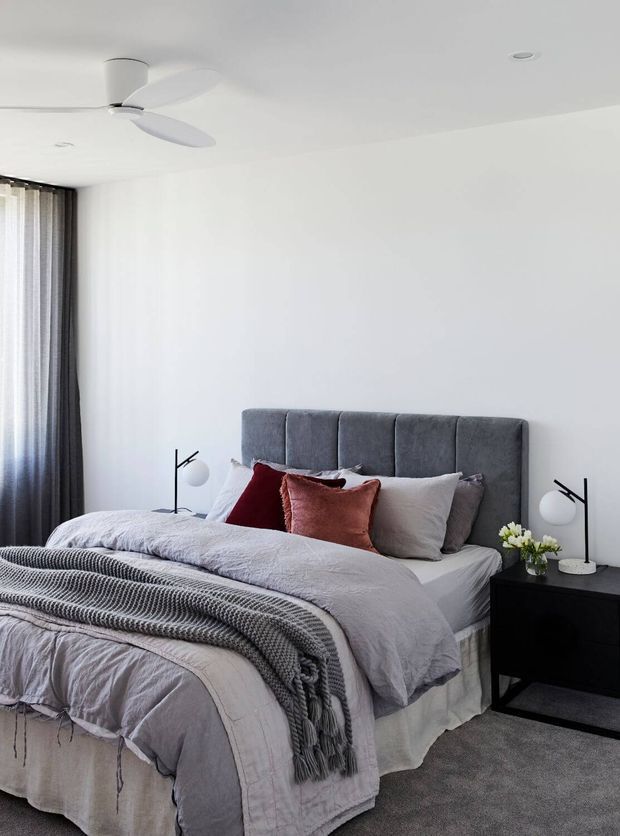 A bright, contemporary bedroom with a grey upholstered vertical-panel headboard, layered grey and white bedding, rust and burgundy accent cushions, black bedside tables, minimalist black-and-white lamps, sheer and dark curtains, and a white ceiling fan. The room has a clean, restrained neutral palette with soft textile layering.