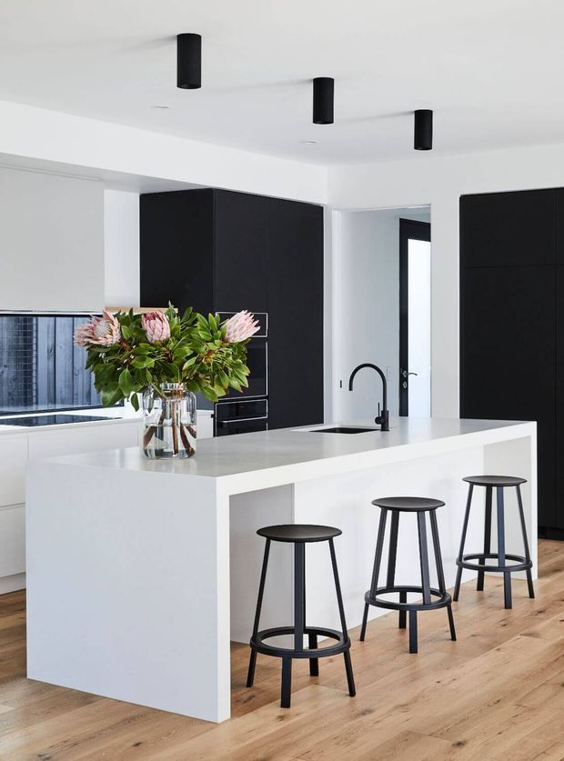 Contemporary minimalist kitchen featuring a large white island with an integrated sink and curved black tap, three black bar stools, matte black floor-to-ceiling cabinetry, white walls and ceiling, black cylindrical ceiling lights, and light timber flooring. A large floral arrangement provides a natural accent.