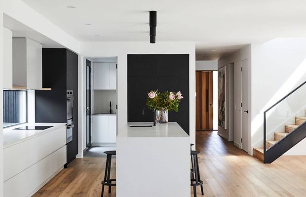Bright contemporary open-plan kitchen and circulation area with a white island, black feature wall, integrated dark cabinetry, timber flooring, and a timber staircase with black metal balustrade. A hallway and utility area extend beyond the kitchen.