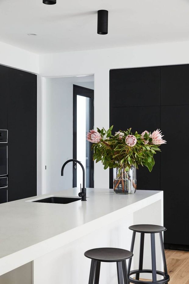 A bright, minimalist kitchen featuring a large white island with an integrated black sink and curved black tap. Matte black floor-to-ceiling cabinetry provides strong contrast against white walls and ceiling, while two simple black stools and a vase of flowers sit on the island. Light timber flooring and black cylindrical pendant lights complete the space.