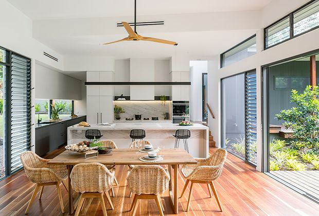 Bright open-plan kitchen and dining area with white built-in cabinetry, a marble-look splashback, central island with stools, and a timber dining table surrounded by woven rattan chairs. Full-height glazing and louvred windows bring in abundant natural light, while polished timber flooring and a sculptural timber ceiling fan add warmth.