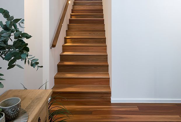 Interior view of a timber staircase rising through a narrow stairwell, with warm wood treads, risers and handrail against white walls. Matching timber flooring extends into the adjoining area, with a wooden console and indoor plants at the base. The space is simple, clean and warmly finished.