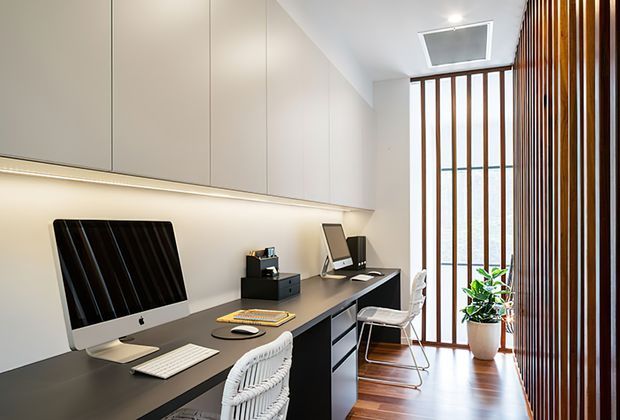 A bright, compact home office with a long built-in dark desk and integrated storage beneath pale full-height cabinetry. Two computer workstations face the wall, while a woven chair and a transparent visitor chair sit along the desk. Warm timber flooring and a vertical timber slat screen frame the glazed end of the room, with a potted plant adding greenery. A ceiling skylight and concealed linear lighting provide additional illumination.