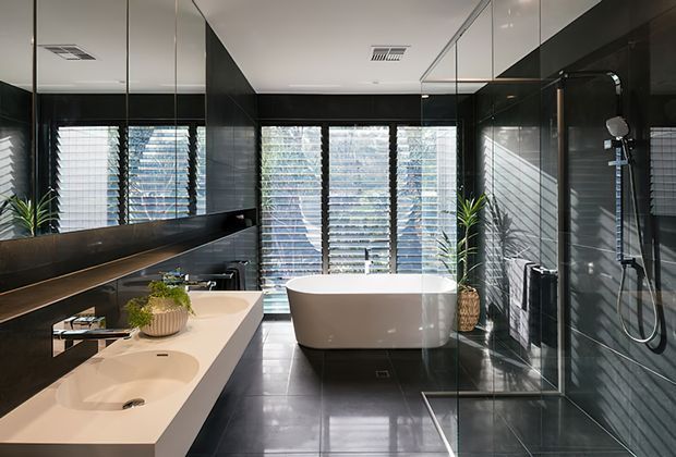 A contemporary, high-contrast bathroom with a freestanding white bathtub positioned in front of louvred windows, a double vanity with a pale stone benchtop, extensive dark wall finishes, mirrored cabinetry, and a frameless glass shower enclosure. The polished dark floor and black fixtures create a sleek, dramatic atmosphere.