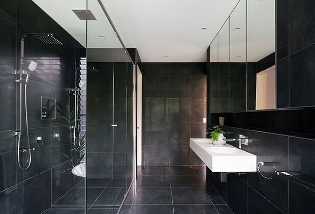 Contemporary dark-toned bathroom with large-format charcoal wall and floor tiles, a frameless glass walk-in shower, wall-mounted black cabinetry, and a long white double vanity with integrated basins. Mirrored cabinetry and reflective surfaces enhance the sleek, spacious appearance.