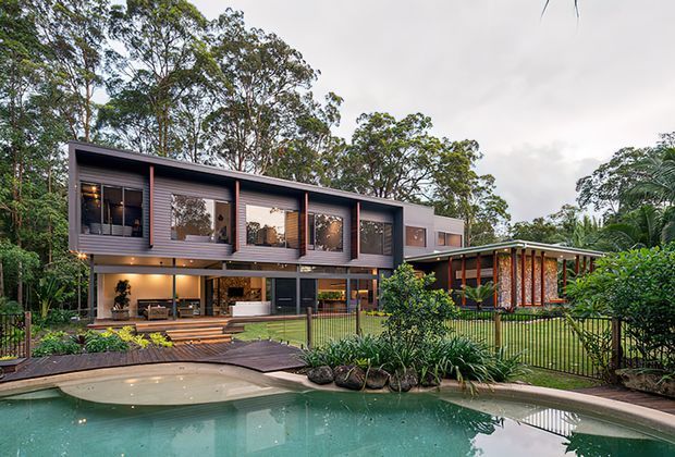A contemporary multi-level home set within lush mature vegetation, viewed from across a curved swimming pool. The dark horizontal facade features extensive glazing, warm timber-toned vertical accents, covered outdoor living areas, and a connected single-storey wing. Landscaped gardens, timber decking, and poolside planting frame the residence.