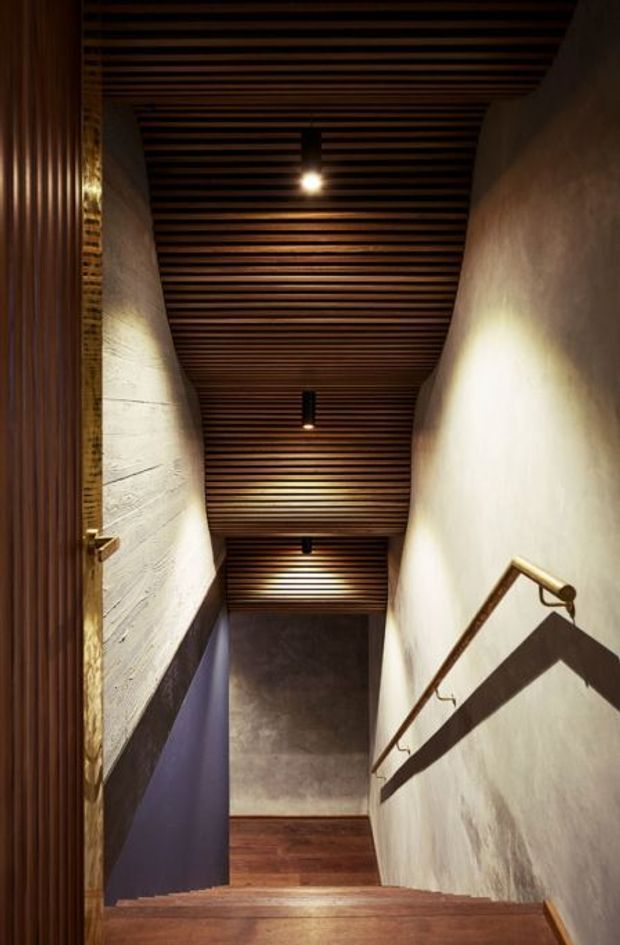 A narrow interior stairway descends through a dramatically lit corridor. Warm timber stairs and a slatted timber ceiling contrast with textured plaster or concrete walls, while slim metal handrails run along the right wall. Small recessed or pendant lights create strong highlights and shadows, producing a dark, refined, material-focused atmosphere.
