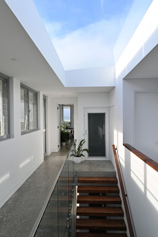 Bright modern interior view of a stairway and upper-level hallway. Floating dark timber stair treads descend beside a clear glass balustrade, with white walls, polished grey concrete or terrazzo-like flooring, extensive glazing, and a large overhead skylight or roof opening bringing in daylight. A glimpse of blue water or sky is visible through the distant opening.