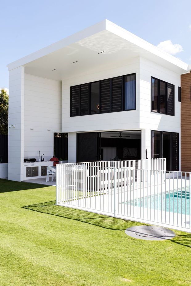 Bright exterior view of a contemporary two-storey white-clad house with a broad flat roof and deep covered terrace. Black-framed louvred windows and large glazed openings contrast with the white horizontal wall cladding. A fenced swimming pool occupies the foreground, with a compact outdoor kitchen and grill area beneath the covered section. The property has a manicured lawn and a clean, geometric design.