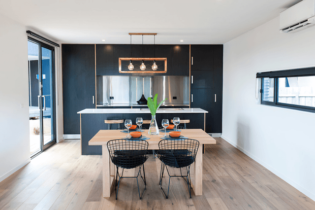 Bright contemporary open-plan kitchen and dining area with a black full-height kitchen wall, reflective splashback, central island with white benchtop, and a light timber dining table paired with black wire-frame chairs. Large glazed doors and windows provide abundant daylight, while pendant lighting and an air-conditioning unit are visible.