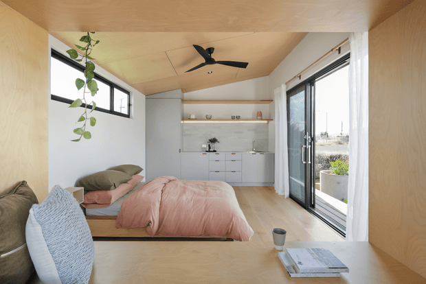 A compact, bright studio-style bedroom with a low platform bed, blush pink bedding, and layered neutral cushions. Pale timber surfaces line the ceiling, walls, and foreground desk or bench, while a minimalist white kitchenette with open shelving and a tiled backsplash sits at the rear. Large sliding glass doors with sheer curtains open to an exterior view, complemented by a high horizontal window, a black ceiling fan, and a hanging trailing plant.