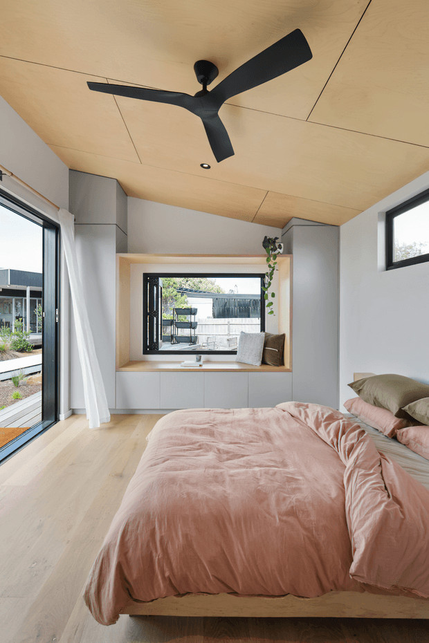 Bright contemporary bedroom with a low timber bed, blush pink bedding, pale timber flooring, and a warm plywood-lined ceiling. Built-in cabinetry surrounds a recessed window seat, while large glazed openings provide abundant natural light. A black three-blade ceiling fan and trailing indoor plant add contrasting details.