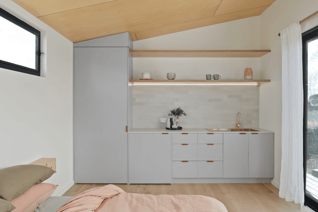 A compact, minimalist bedroom with an integrated kitchenette along the far wall. The room features pale grey flat-panel cabinetry, open timber shelves, a light tiled splashback, a small sink and cooktop, pale timber flooring, a warm timber-lined ceiling, and soft neutral bedding in the foreground. Black-framed windows and sheer white curtains bring in natural light.