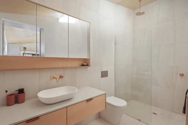 Bright contemporary bathroom with large white wall and floor tiles, a frameless glass shower enclosure, wall-mounted rain shower, freestanding oval basin on a pale timber vanity, and a large mirrored cabinet above a warm timber shelf. A toilet and minimal copper-toned fixtures complete the clean, neutral space.