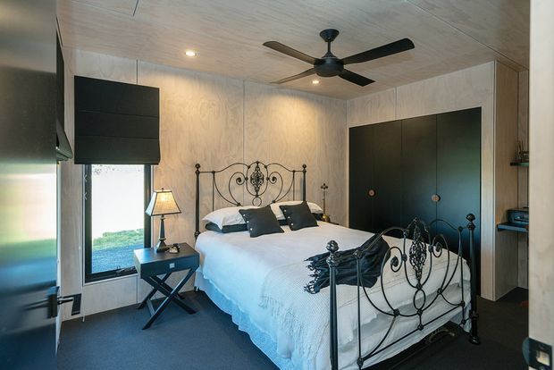 A compact, warmly lit bedroom with a white bed dressed in layered white bedding and dark accent cushions. The room features ornate black wrought-iron bed frames, dark built-in wardrobe cabinetry, pale timber wall and ceiling panelling, dark carpet, a bedside table and lamp, a ceiling fan, and a large window overlooking greenery.