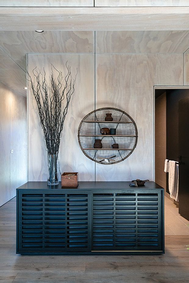 A refined interior vignette featuring a dark slatted timber console against pale plywood wall and ceiling panelling. A circular metal wall shelf displays small pottery objects, while a tall glass vase with dried branches adds height. An open doorway on the right reveals a dark-toned bathroom or dressing area with a towel rail. The space has warm natural materials, restrained styling, and strong vertical and horizontal lines.