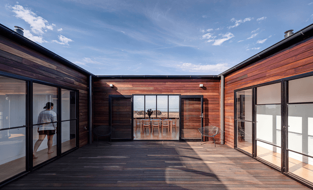 A contemporary single-storey courtyard home arranged around a spacious timber deck. Dark horizontal timber-clad walls and black-framed floor-to-ceiling glazing enclose the courtyard, with a dining area visible through the central opening and a person visible inside the left wing. The open sky and warm timber surfaces create a calm indoor-outdoor architectural setting.