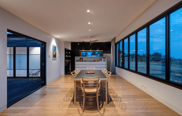 A long, contemporary dining room with a timber floor, large dark-framed windows overlooking a blue-toned landscape, and full-height sliding glazing opening to an exterior deck. A rectangular dining table with wooden chairs occupies the centre, while a dark kitchen or bar joinery area with an illuminated aquarium-like feature sits at the far end. Recessed ceiling lights provide a clean, refined finish.