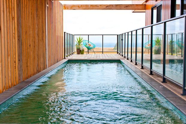 A narrow rectangular swimming pool occupies a sheltered outdoor terrace or courtyard, bordered by timber decking, warm vertical timber cladding, and black-framed glass balustrades. The pool leads toward an open ocean view, with tropical potted plants and a colourful outdoor chair at the far end. The space is bright, contemporary, and coastal in character.