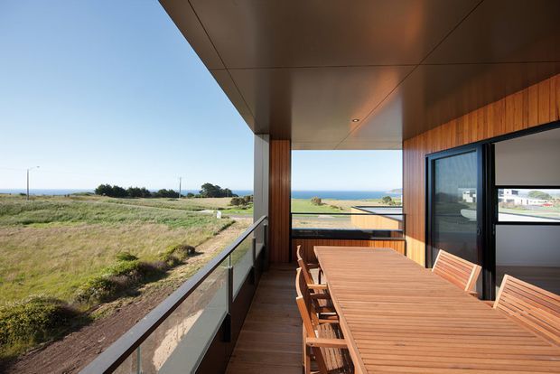 A covered outdoor dining terrace with a large timber dining table and matching chairs, framed by warm vertical timber cladding and dark-framed glazing. The terrace overlooks a grassy coastal landscape and the ocean beyond, with a glass balustrade and recessed ceiling lighting creating a sheltered, contemporary entertaining space.