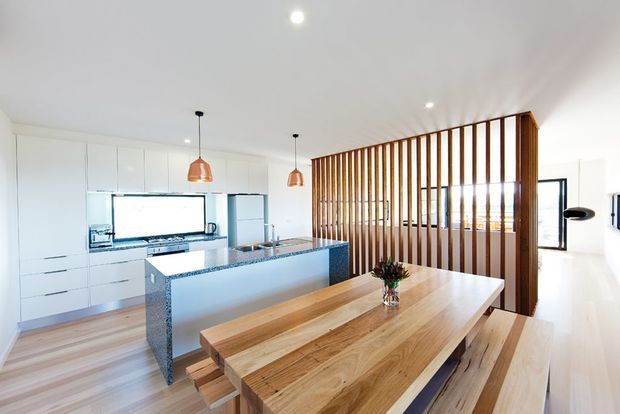 Bright open-plan kitchen and dining area with extensive white built-in cabinetry, a long stone island, timber flooring, and a substantial natural timber dining table with benches. A vertical timber slatted screen separates the dining zone from the adjacent living area, while pendant lights and recessed downlights provide illumination.
