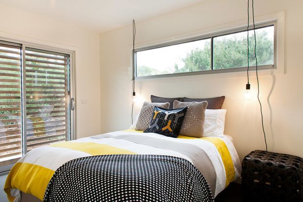 A bright, compact modern bedroom with a low-profile bed dressed in white and yellow bedding, patterned cushions, and a black-and-white textured throw. Full-height louvred glazing and a wide horizontal window bring in natural light and views of greenery, while exposed pendant bulbs hang beside the bed. A dark patterned ottoman sits at the foot of the bed.