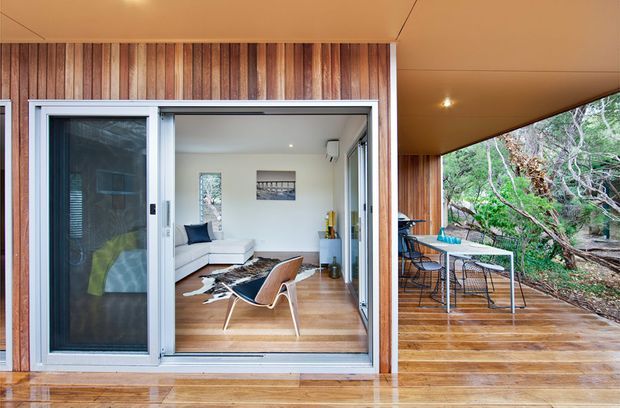 A compact modern timber-clad dwelling with large sliding glass doors opening onto a covered timber deck. The view shows a simply furnished living room with a white sectional sofa, leather lounge chair, low rug, and timber flooring, alongside an outdoor dining area integrated into a wooded setting.