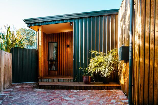 A compact modern dwelling entrance viewed from a small brick-paved courtyard. The facade combines vertical warm timber cladding with dark ribbed metal panels, a deep covered entry, timber-framed glazing, and a low step-up porch. Potted tropical planting and timber privacy fencing frame the entry.