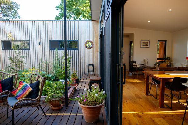 A bright contemporary open-plan interior opening directly onto a timber deck and planted courtyard. The image shows a dining area with a solid timber table and chairs, warm timber flooring, recessed ceiling lights, and large black-framed glazed folding or sliding doors. Outside, the deck includes wicker lounge chairs, potted plants, flowers, and a narrow landscaped courtyard beside a vertically clad wall.