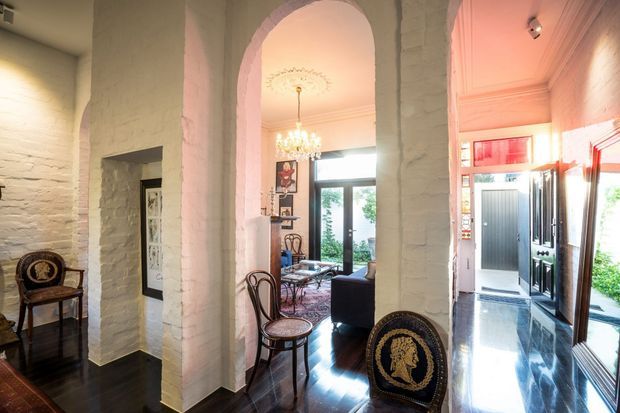 Interior view of a character-filled entry and living area with multiple white-painted brick arched openings, dark polished timber flooring, ornate ceiling detailing, a chandelier, and traditional furniture. The image looks through the entry toward a bright glazed doorway and adjoining sitting space, with a reflective black-and-white tiled entry zone on the right.