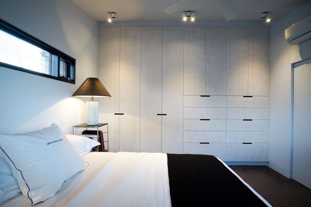 A compact, contemporary bedroom with a neatly made bed, white bedding and a black throw, extensive light wood built-in wardrobe cabinetry, a bedside table with a lamp, a horizontal window with blinds, recessed ceiling lights, and a wall-mounted air conditioner. The room has a calm neutral palette and functional built-in storage.