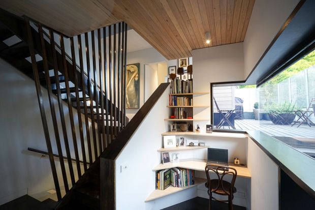 Compact, contemporary interior featuring a dark timber staircase with vertical balustrades, a built-in stepped bookshelf and desk nook, warm timber-lined ceiling, and expansive corner glazing opening toward an outdoor deck.