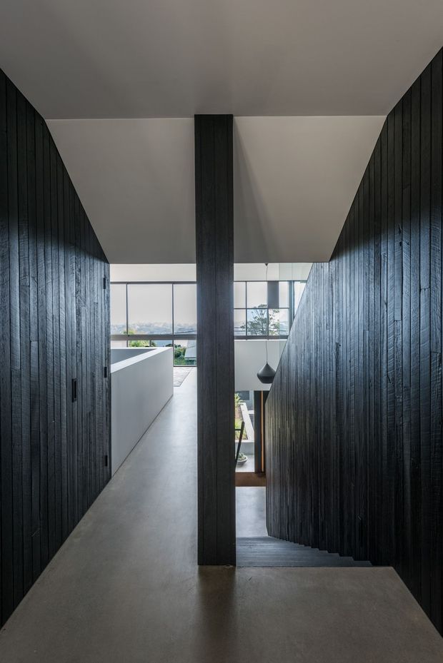 A dramatic interior stairway and circulation corridor with dark vertical timber wall cladding, a central dark timber-clad structural column, and a smooth pale concrete or grey floor. The stair descends beside an open void toward a bright glazed area, with a minimalist black pendant light and glimpses of greenery and an adjacent lower-level space. The composition emphasizes strong linear perspective, contrast between dark timber and light surfaces, and abundant natural light.