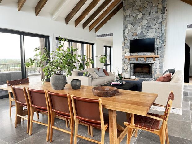 A bright open-plan living and dining area with a long timber dining table and leather-upholstered timber chairs in the foreground. The living zone includes cream sofas, a dark coffee table, and a stone-clad fireplace with a timber mantel and wall-mounted television. Large black-framed windows open to a balcony or view, while exposed timber beams run across the high ceiling. The space has a warm, contemporary rustic character.