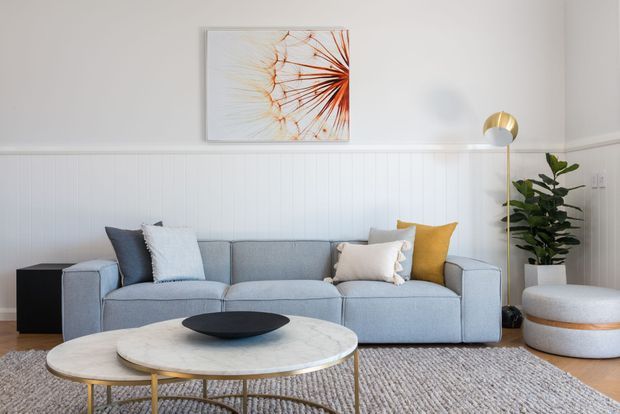 Bright, inviting living room with a large pale blue-grey sectional sofa, layered neutral cushions, white vertical wall panelling, a light timber floor, textured area rug, and nested round coffee tables with white stone-like tops and slim gold-toned frames. A sculptural floor lamp, potted plant, and framed botanical artwork add warmth and visual interest.