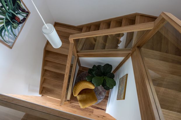 Interior view looking down through a timber staircase, showing warm wood stair treads, a geometric timber balustrade, white walls, a hanging cylindrical pendant light, and a small seating area with a mustard velvet chair and plant.