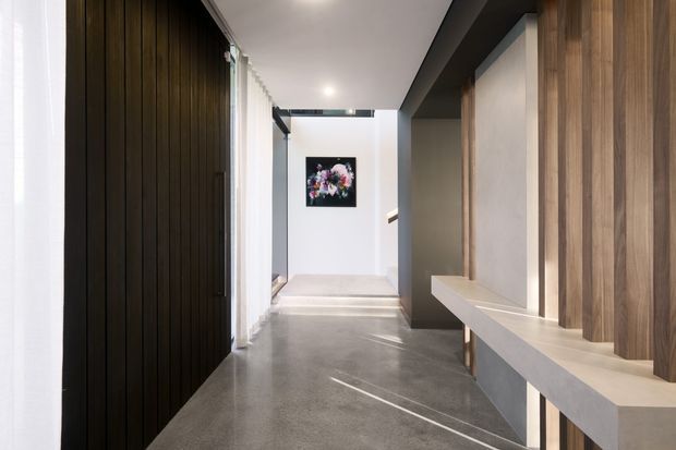 A modern interior hallway with a polished grey concrete floor, dark vertical timber wall panelling on the left, and a built-in light stone or concrete console on the right framed by warm timber battens. Recessed ceiling lighting, sheer curtains, and a floral artwork at the end of the corridor create a refined, minimalist composition.