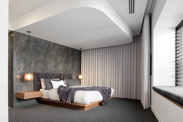 A contemporary bedroom with a low floating timber bed, upholstered headboard, layered grey and white bedding, integrated bedside tables, pendant lights, a dramatic textured grey feature wall, full-height curtains, and a window seat with horizontal blinds. The room has a sculptural curved white ceiling bulkhead and dark carpet flooring.
