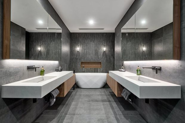 A symmetrical, contemporary bathroom with twin floating white vanities, integrated rectangular basins, wall-mounted black tapware, large mirrored walls, grey stone-look wall and floor finishes, dark vertical feature panelling, timber vanity cabinetry, and a freestanding sculptural bathtub centered at the rear.