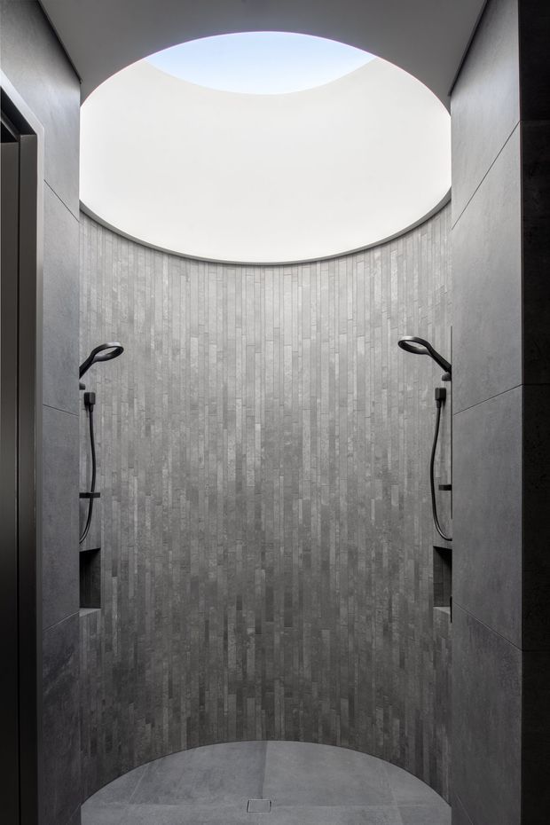 A dark, minimalist walk-in shower enclosure with dual wall-mounted handheld shower fittings facing each other. The curved rear wall is clad in narrow vertical grey stone-look tiles, with large-format grey wall panels on the side walls and a matching grey floor. A large circular skylight or oculus creates a bright overhead opening and curved ceiling detail.